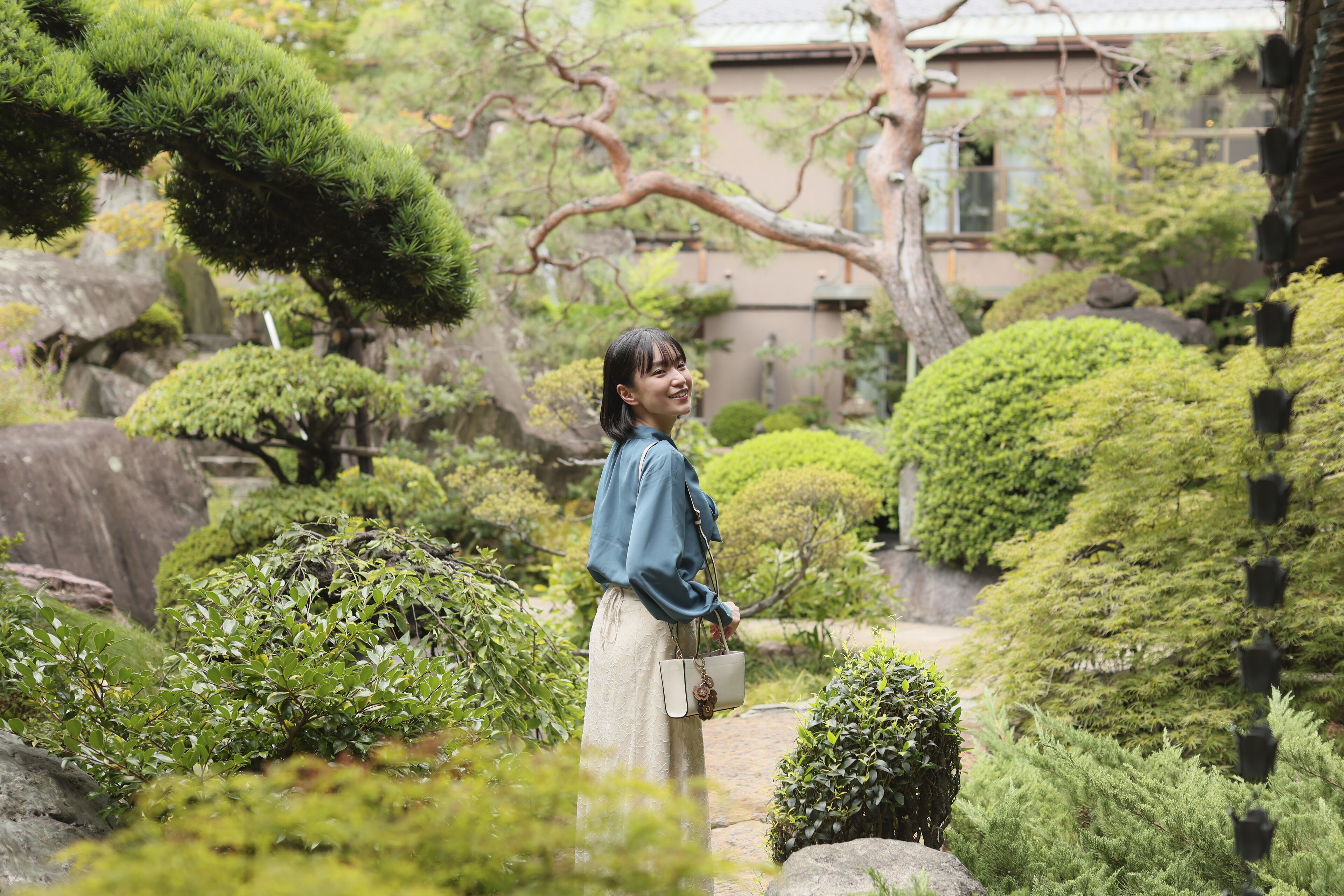 A woman in a garden with various types of greenery and a building in the background.