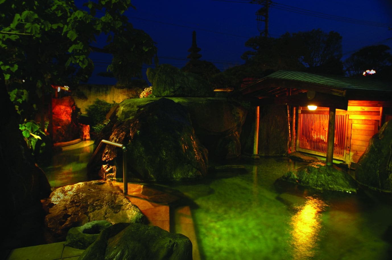 Vista nocturna de un jardín japonés con una formación rocosa, un pequeño estanque y una edificación tradicional de madera con techo verde.