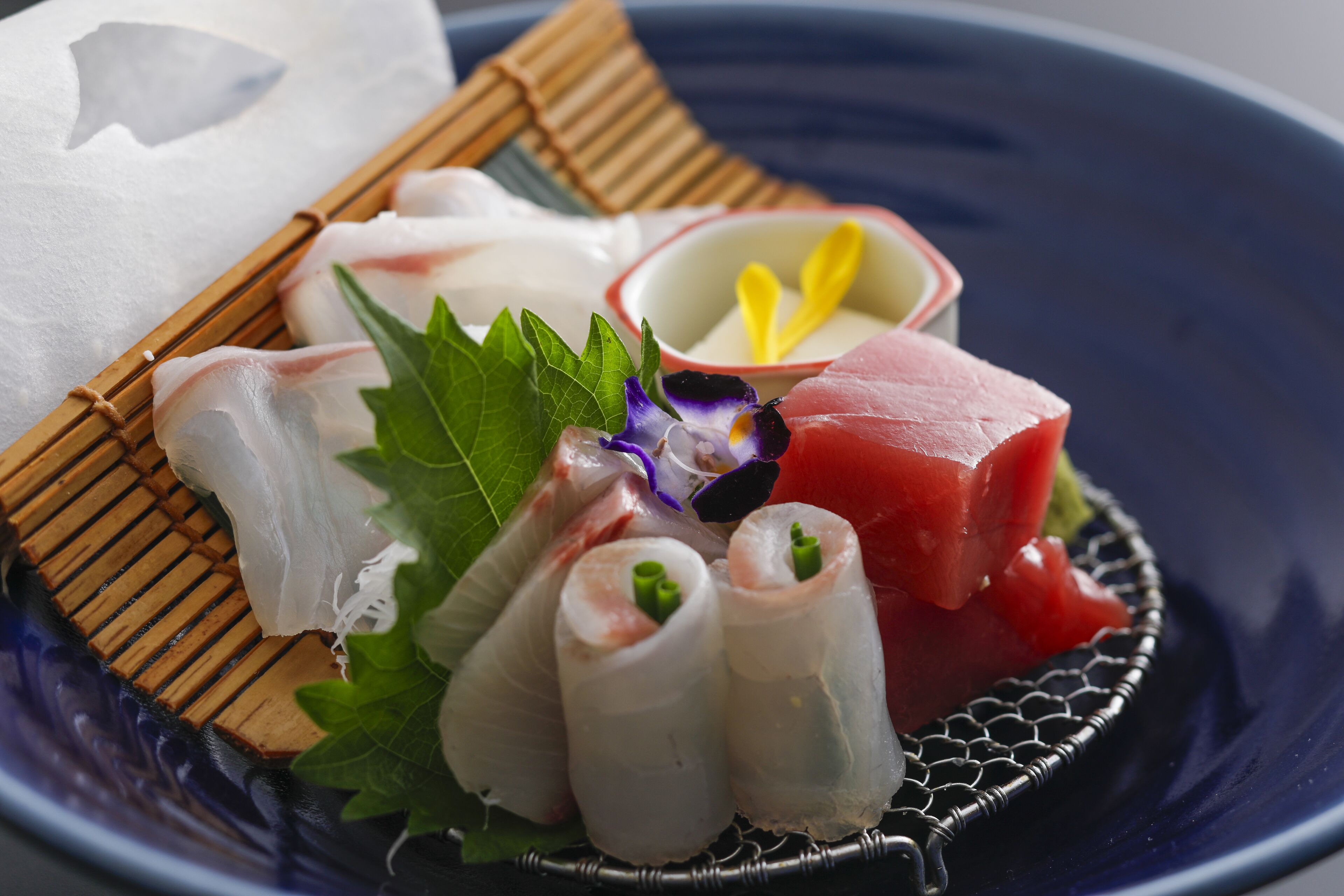 A plate of sashimi with various types of fish, including a piece of raw tuna, on a blue plate with a bamboo mat and a small bowl of wasabi.