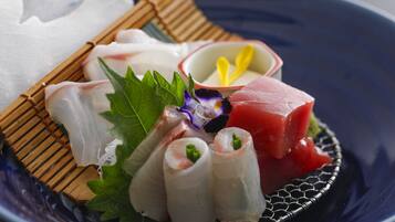 A plate of sashimi with various types of fish, including a piece of raw tuna, on a blue plate with a bamboo mat and a small bowl of wasabi.