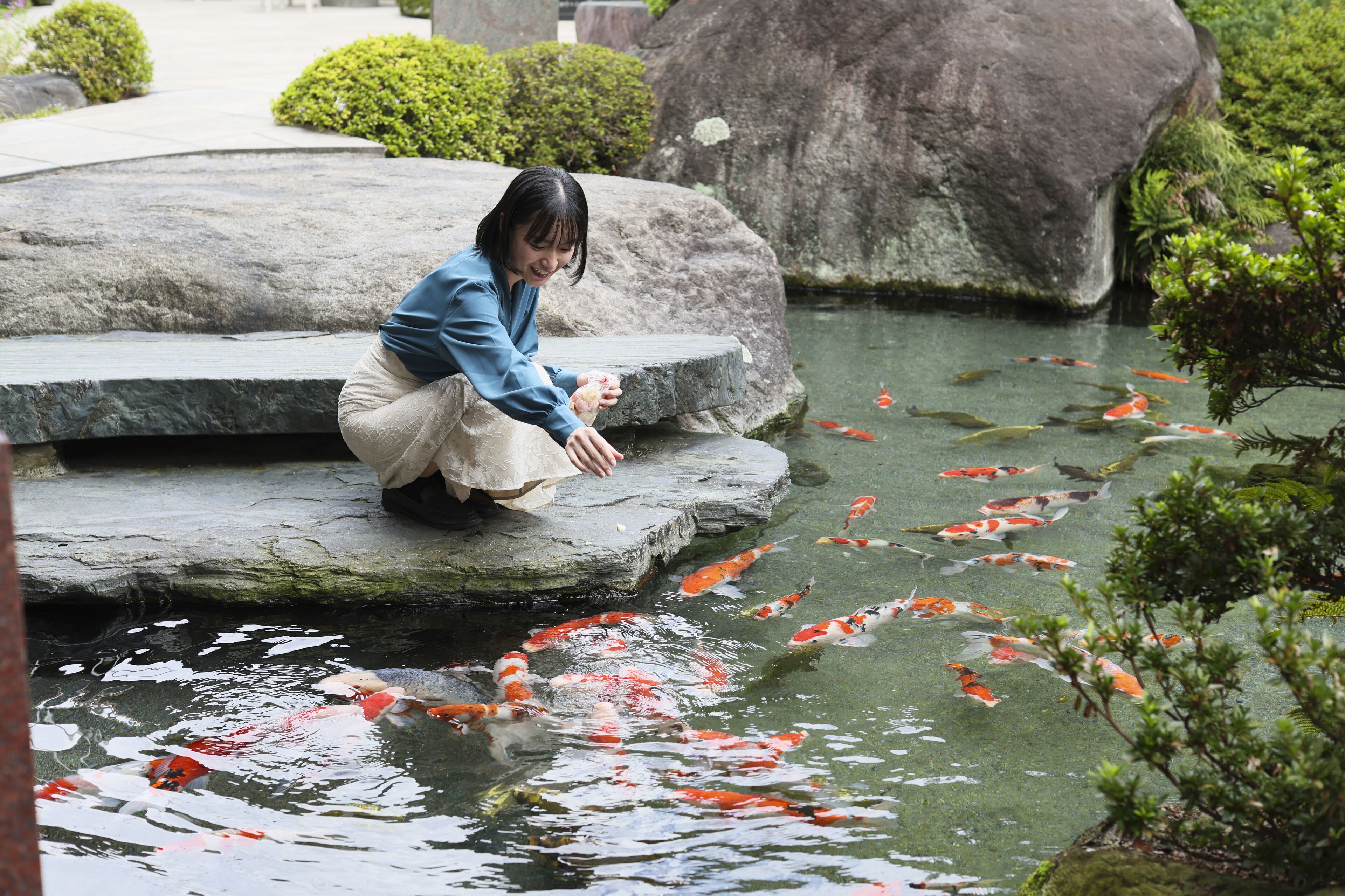 A person sitting on a stone ledge by a koi pond with large rocks and greenery in the background.