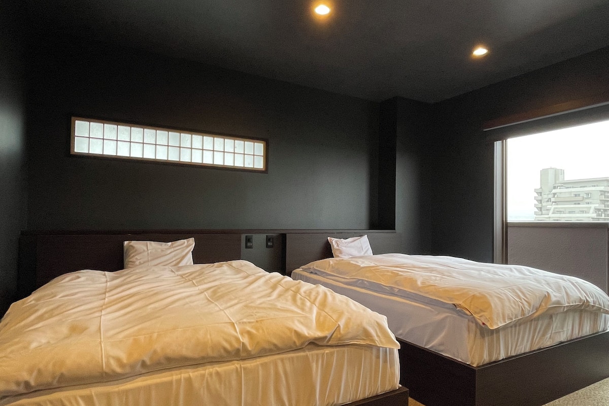 Une chambre d’hôtel avec deux lits, une fenêtre donnant sur des immeubles et un luminaire fixé au mur.