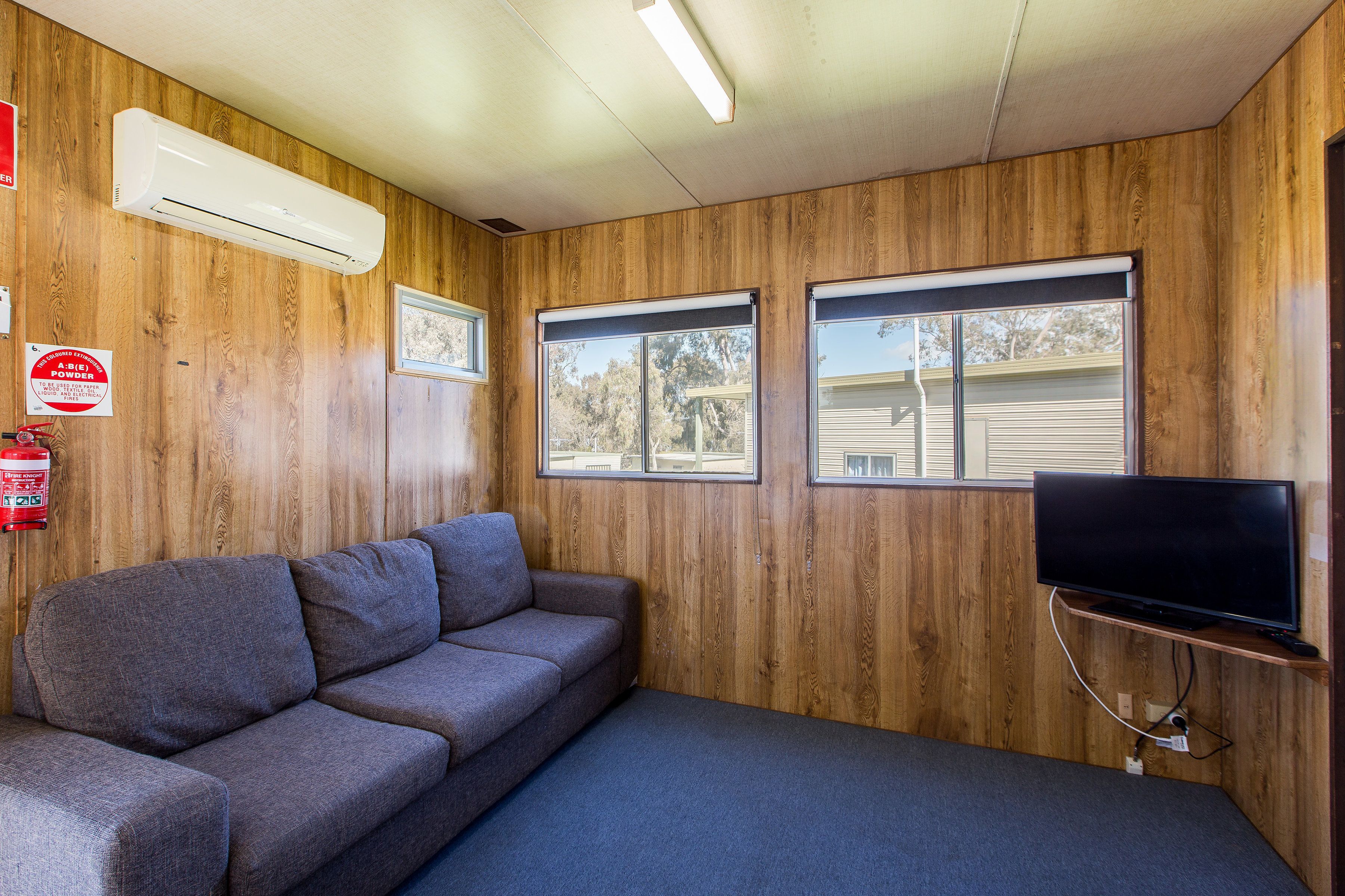 A room with wooden walls, a gray sofa, a flat-screen TV on a stand, and two windows with blinds.