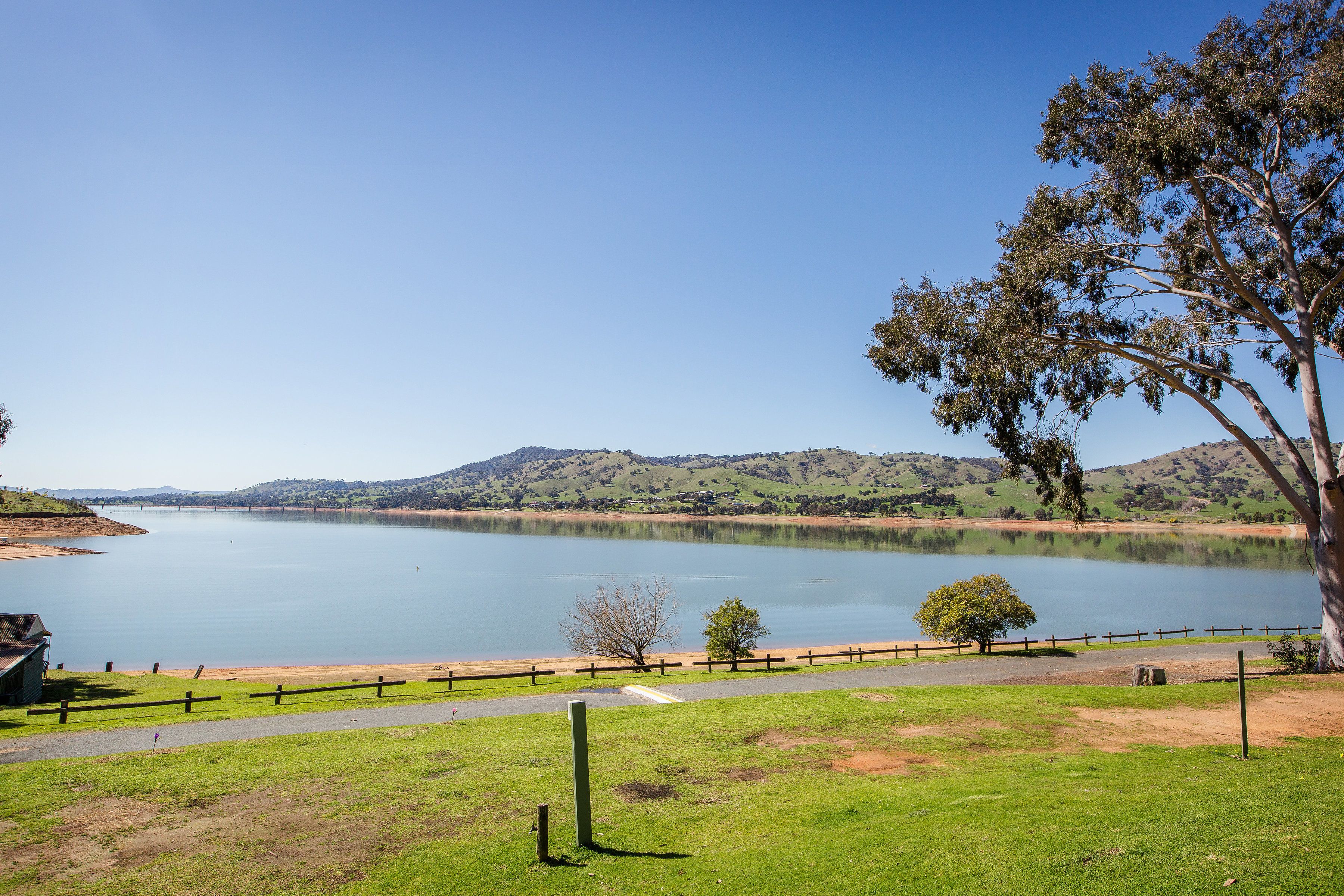 A lakeside view with a wooden fence, a paved path, and a grassy area.