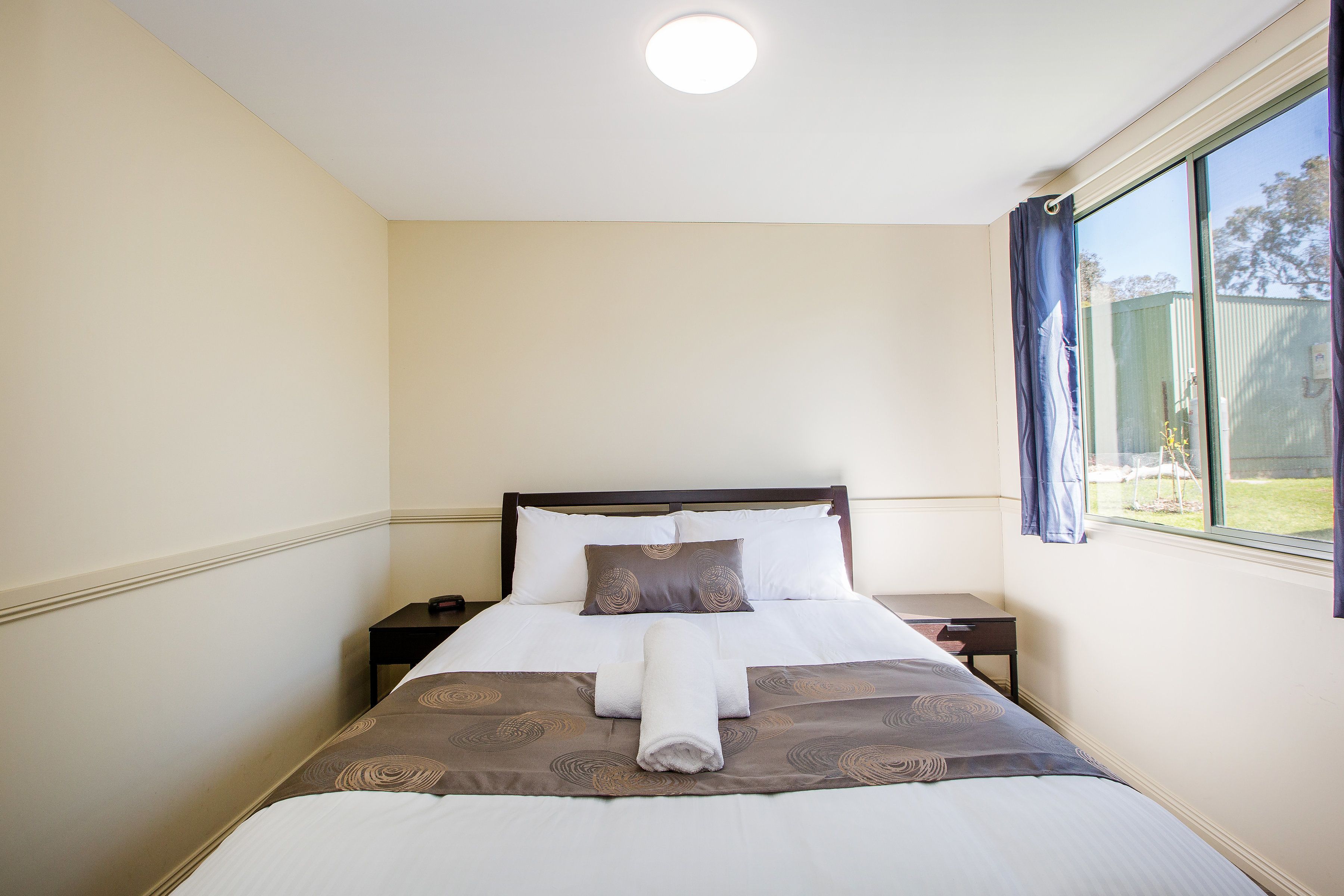 A neatly made bed with a brown patterned bedspread and white linens, a bedside table with a lamp, and a window with curtains.