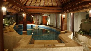 Una piscina interior con techo de paja, escalones de piedra y una zona de estar con vistas a la piscina.