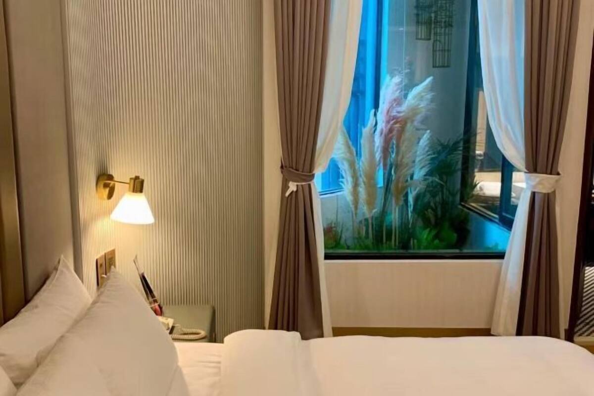 Hotelkamer met een bed, nachtkastje, lamp, gordijnen en een raam met uitzicht op beplanting.