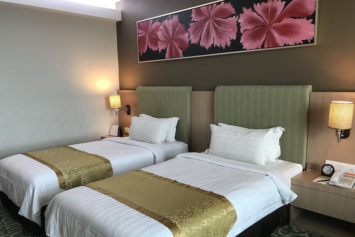 A hotel room with two beds, each with white linens and a gold accent. There is a large floral artwork above the beds, a nightstand with a lamp, and a clock on the wall.
