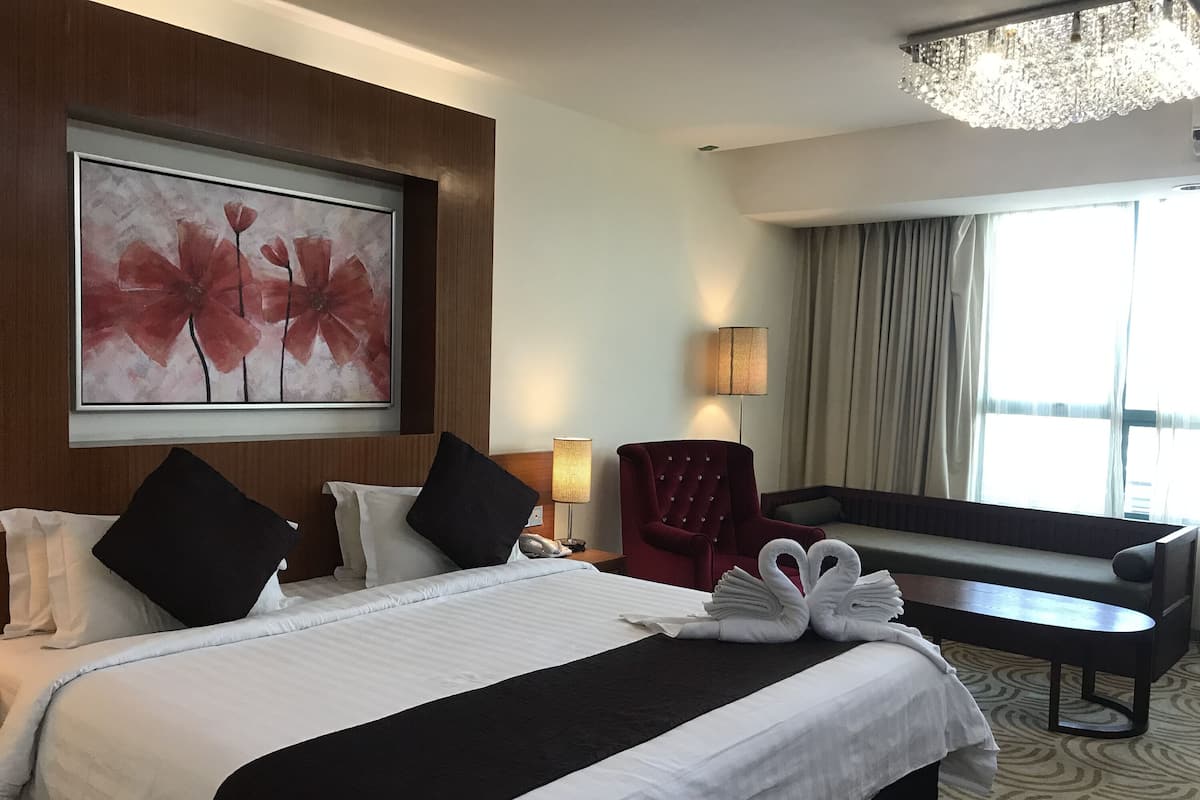 A hotel room with a large bed, a red armchair, a sofa, a chandelier, and a painting of flowers.