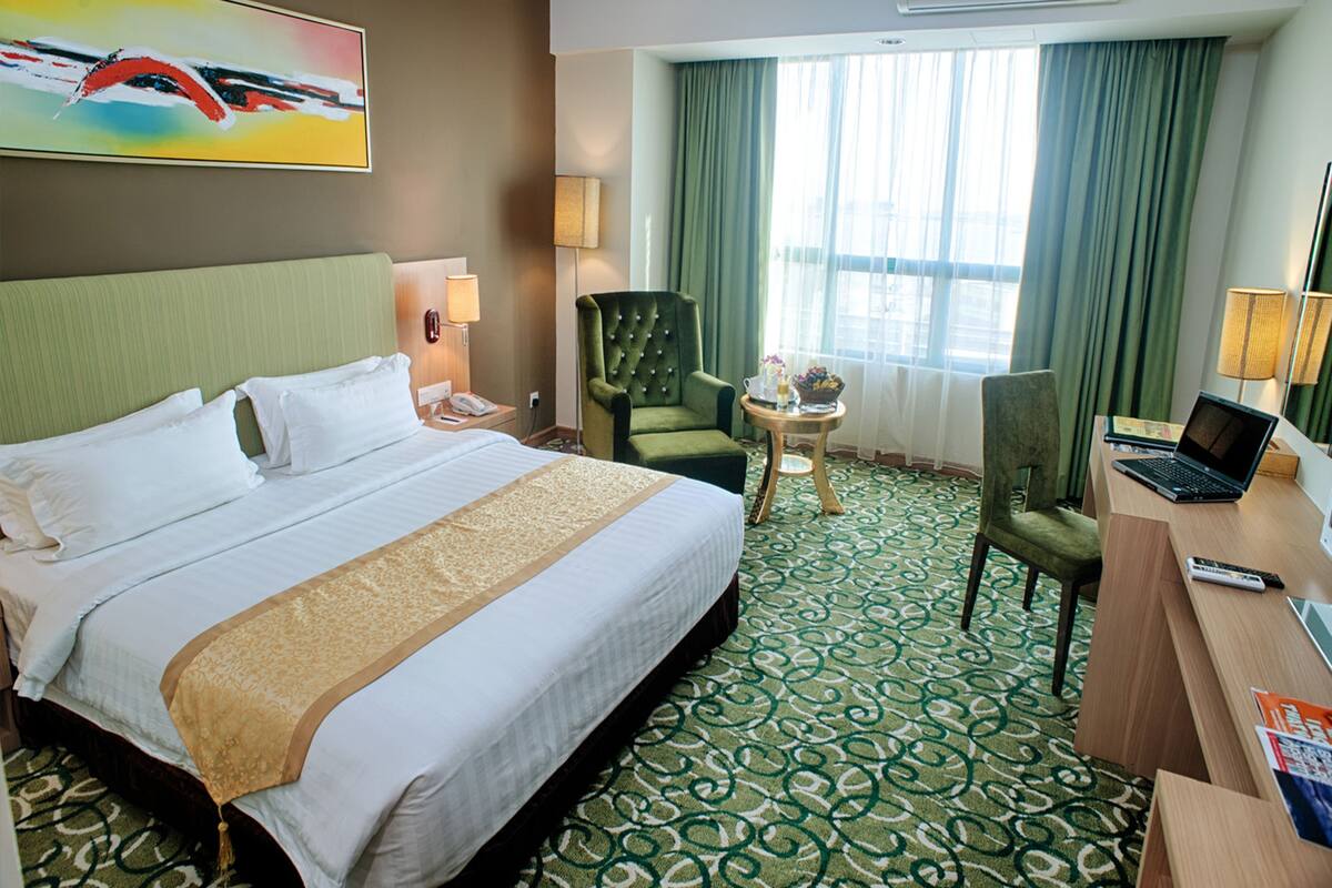 A hotel room with a large bed, a desk with a laptop, a green armchair, and a small table with a lamp and a vase.
