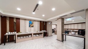 Un moderno vestíbulo de hotel con un mostrador de recepción, una zona de buffet con platos ordenados y una cocina visible al fondo.