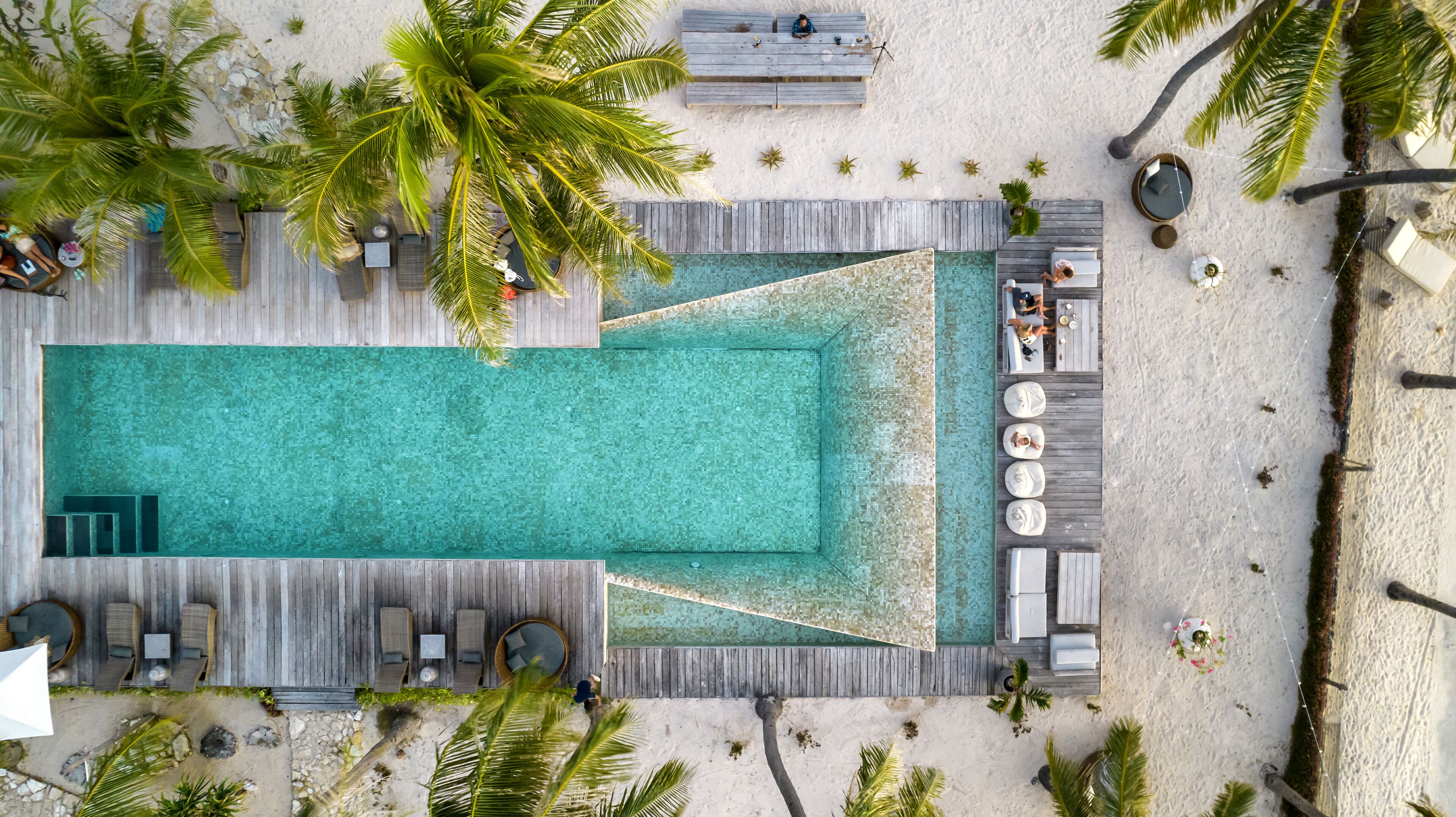 An aerial view of a resort with a central swimming pool, wooden deck seating, and palm trees.