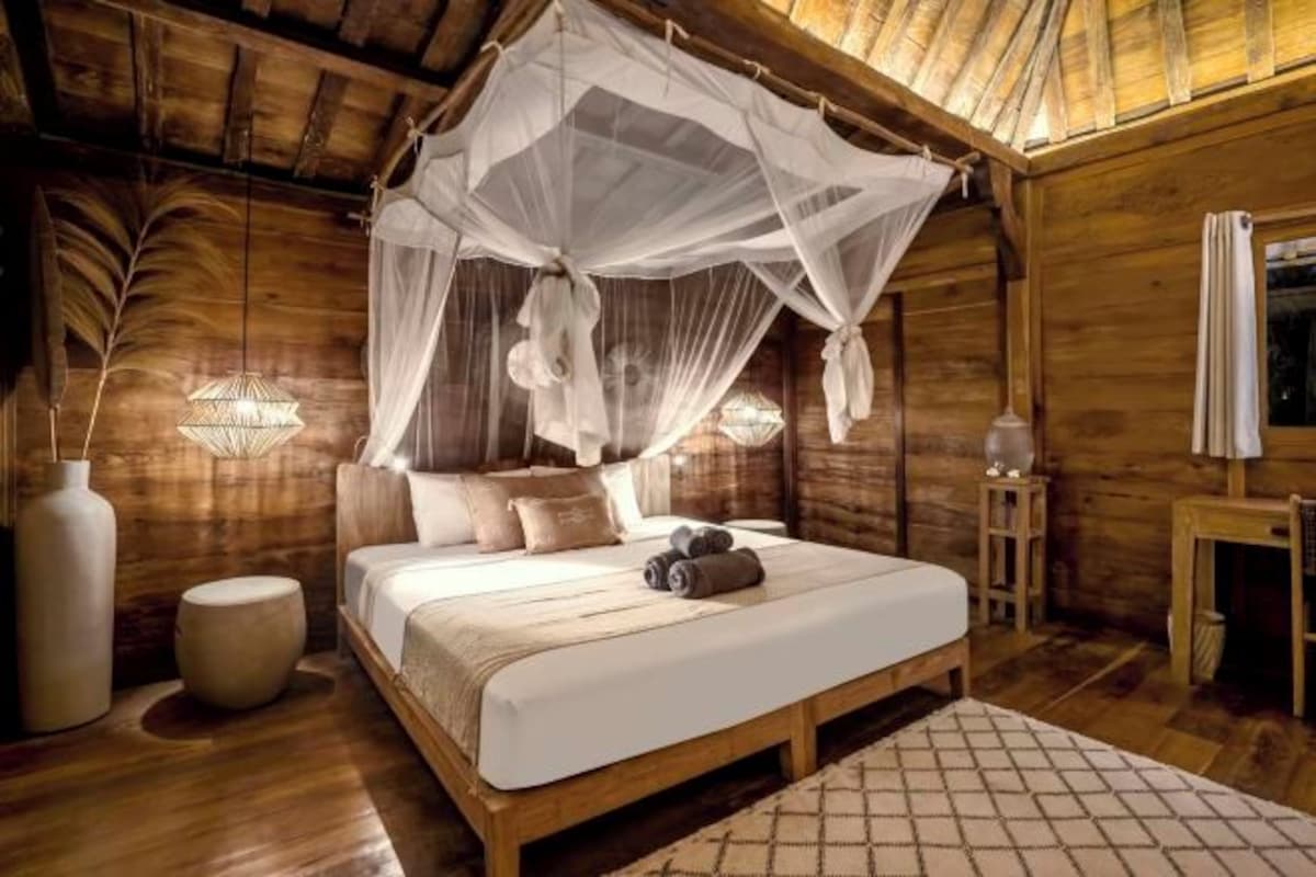 A wooden room with a bed, a canopy, a vase, and a lamp.