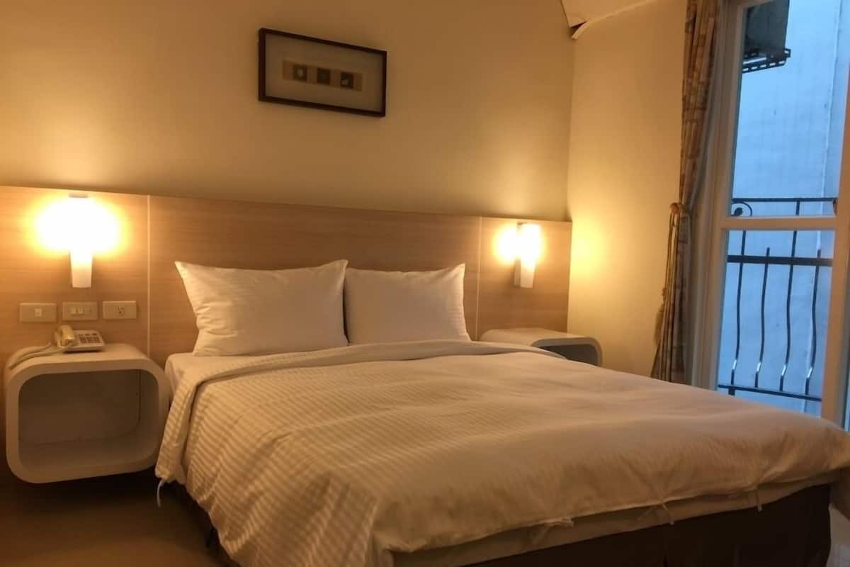 A hotel room with a large bed, two bedside tables, a nightstand, a wall-mounted light, and a window with a view.