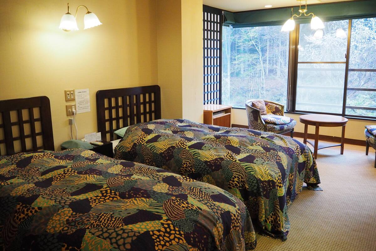A hotel room with two beds, a window with a view of trees, a chair, and a small table.