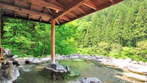 Un pavillon en bois surplombe une source d’eau chaude naturelle, entourée de rochers et d’une végétation luxuriante.
