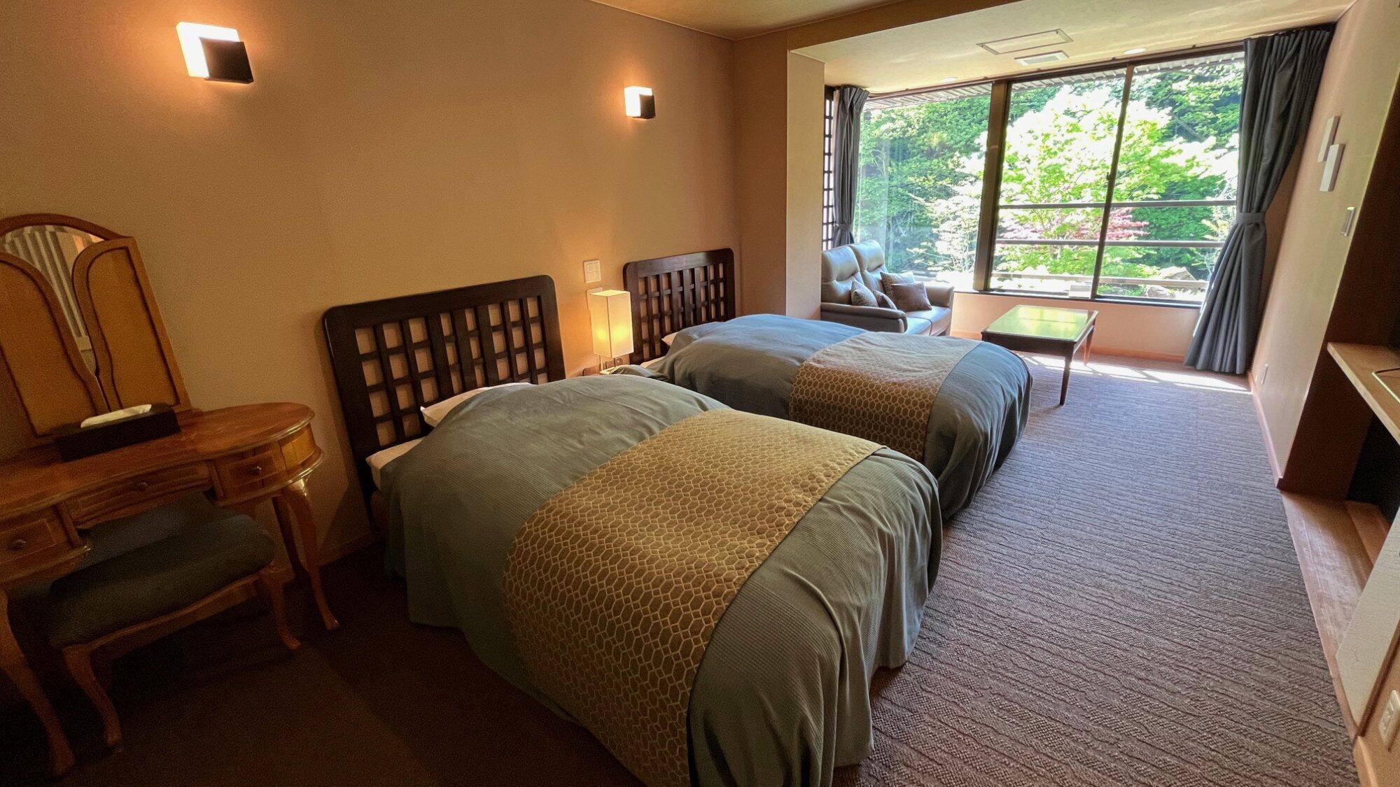 A hotel room with two beds, a desk, a chair, a lamp, and a window with a view of trees.