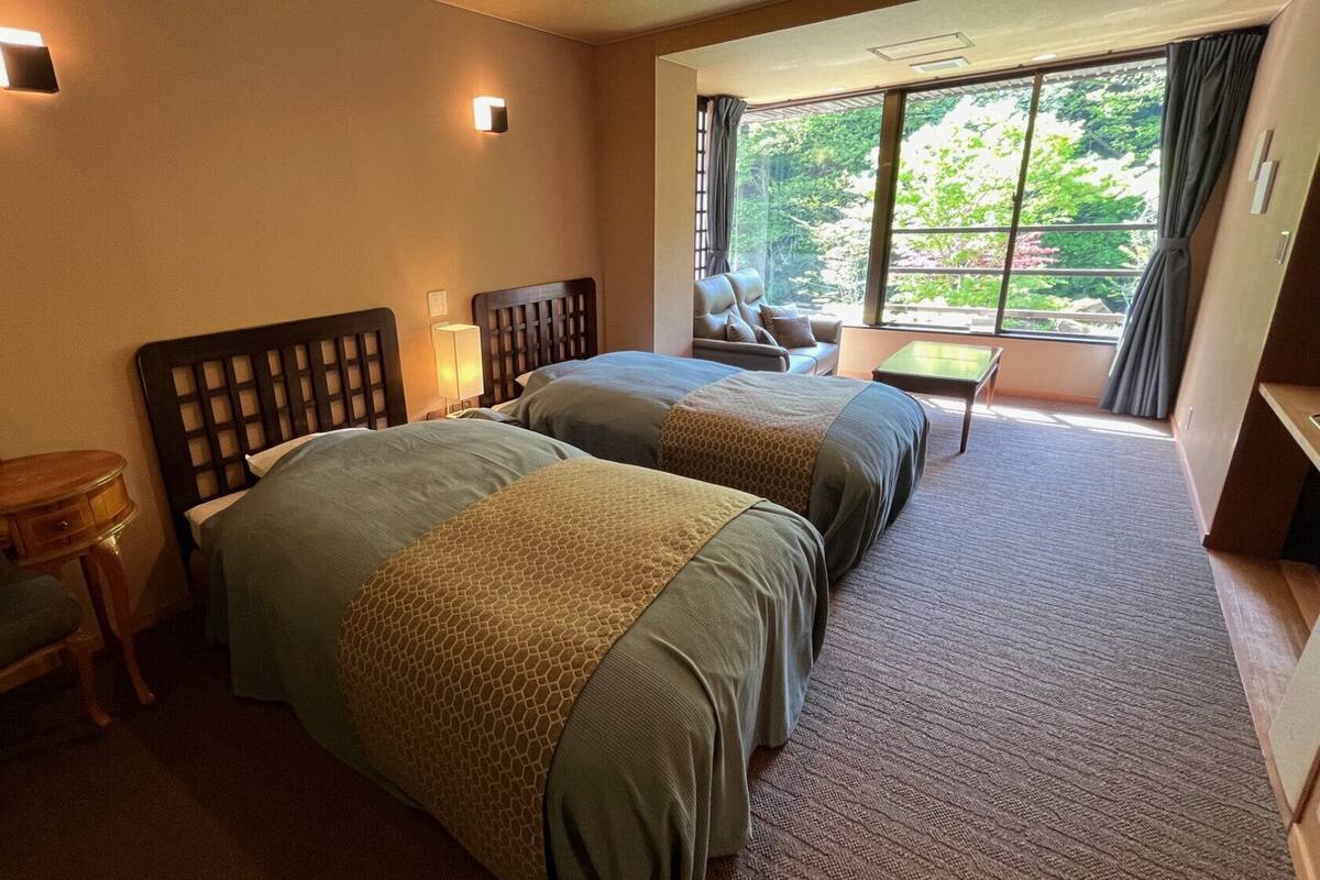 A hotel room with two beds, a desk, a chair, a lamp, and a window with a view of trees.