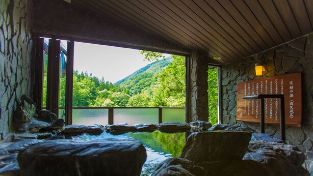 Un espace de sources chaudes doté de murs en pierre et d’un plafond en bois, un mur en pierre orné d’inscriptions en japonais, et une vue sur un lac et une forêt.