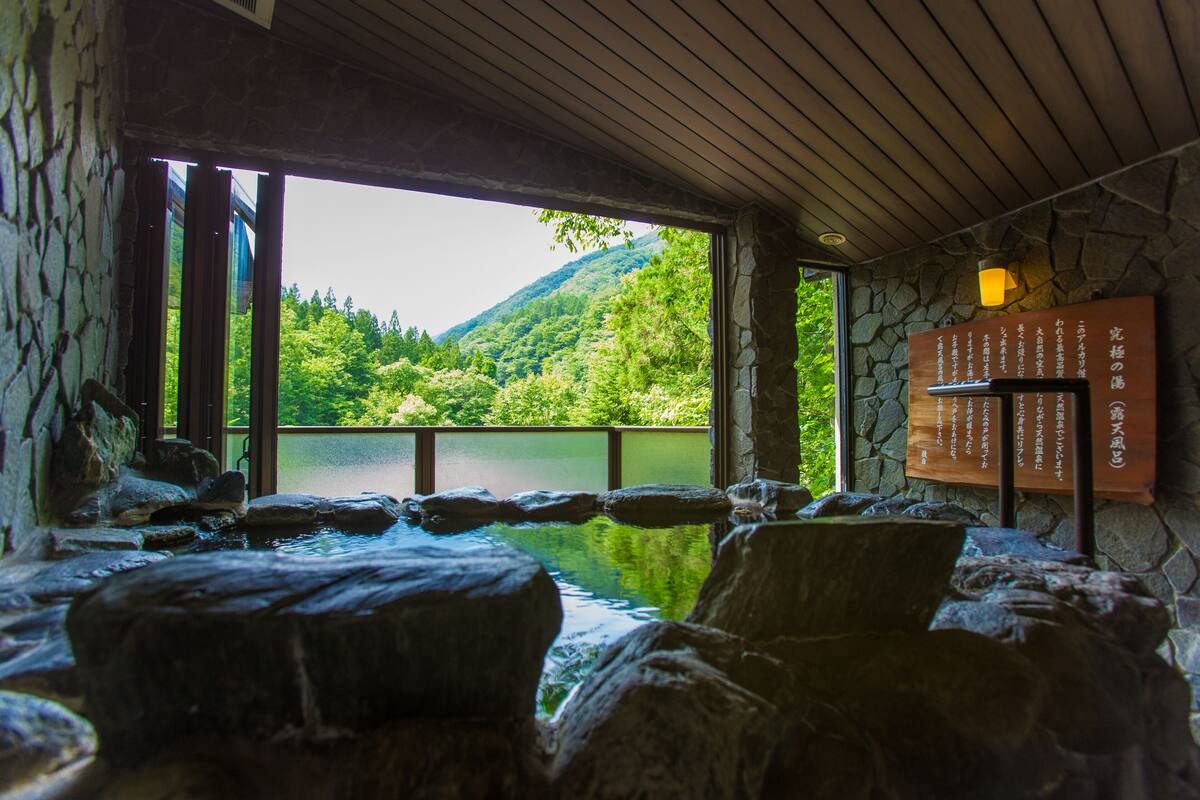 A hot spring area with stone walls and a wooden ceiling, a stone wall with Japanese text, and a view of a lake and forest.