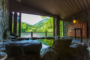 Un área de aguas termales con paredes de piedra y techo de madera, una pared de piedra con texto japonés, y vistas a un lago y bosque.