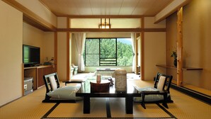 Une pièce de style japonais traditionnel, avec un sol recouvert de tatamis, une petite table basse avec des coussins, un téléviseur et une grande fenêtre donnant sur un paysage verdoyant.