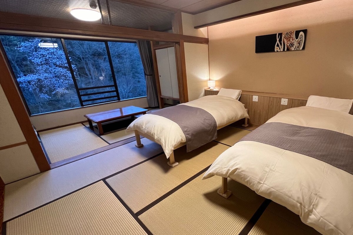 A traditional Japanese-style room with tatami flooring, two beds, a small table, and a window with a view of trees.