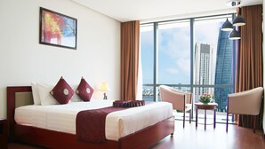 Habitación de hotel con una cama grande, un escritorio, una silla y vistas a edificios altos a través de ventanas amplias.