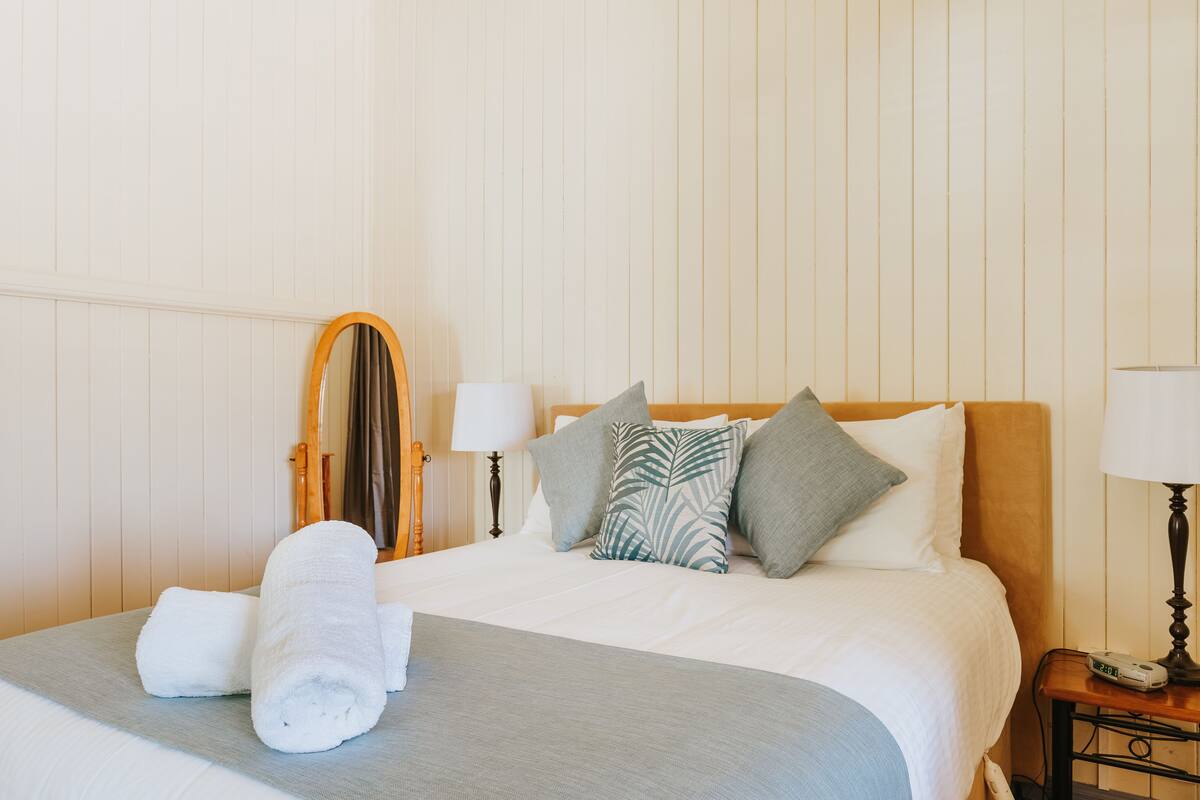 A neatly made bed with white linens, a grey throw blanket, and a set of white towels. There are pillows with various patterns, including a palm leaf design. A wooden-framed mirror hangs on the wall, and there are two bedside lamps on either side of the bed.