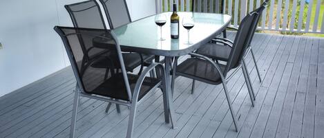 A glass-top table with four chairs, a bottle of wine, and two glasses on a wooden deck.