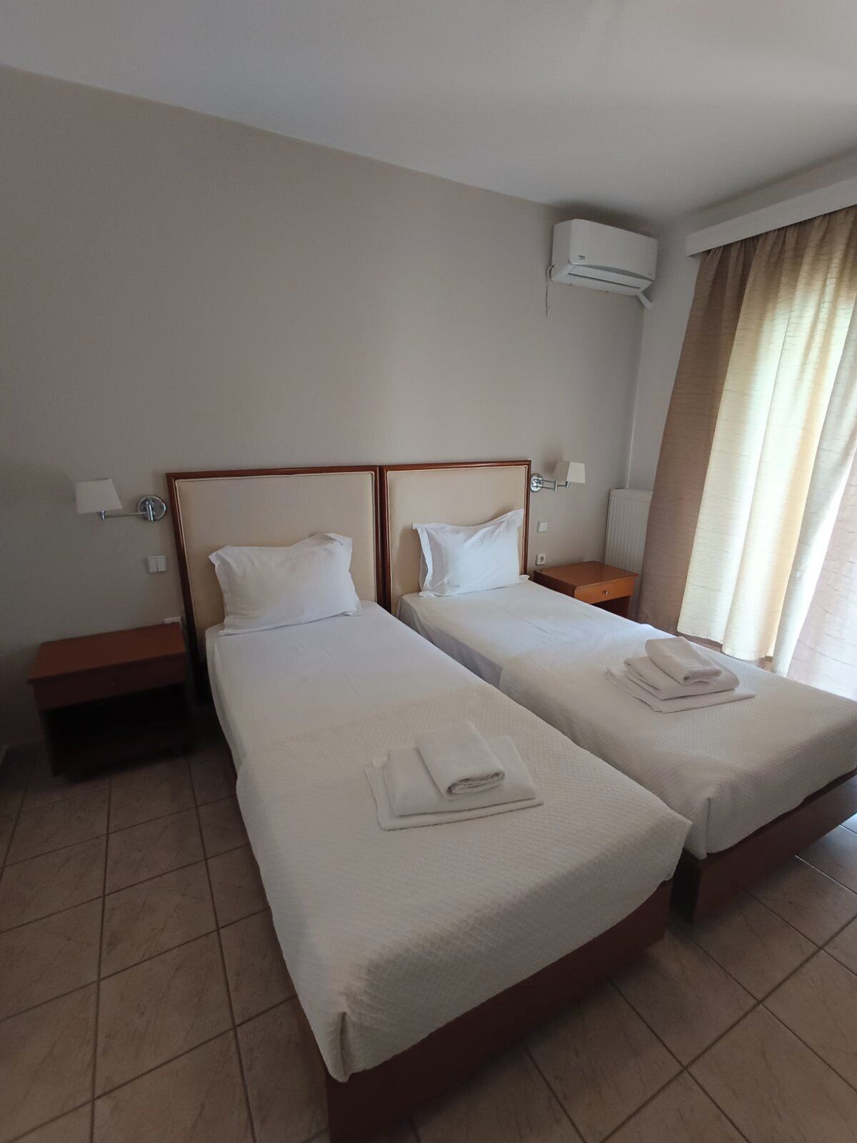 A hotel room with two beds, a nightstand, a lamp, an air conditioning unit, and a window with curtains.