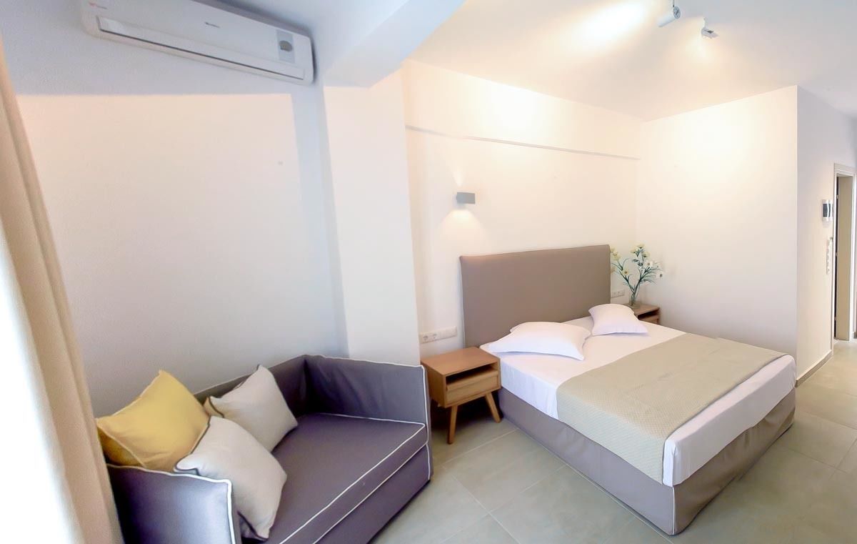 A compact hotel room with a bed, a small sofa, a nightstand, and a wall-mounted air conditioning unit.