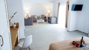 A modern hotel room with a sofa set, a dining table with a fruit platter, a TV mounted on the wall, and a lamp. - Stavros Beach Hotel (Volvi)