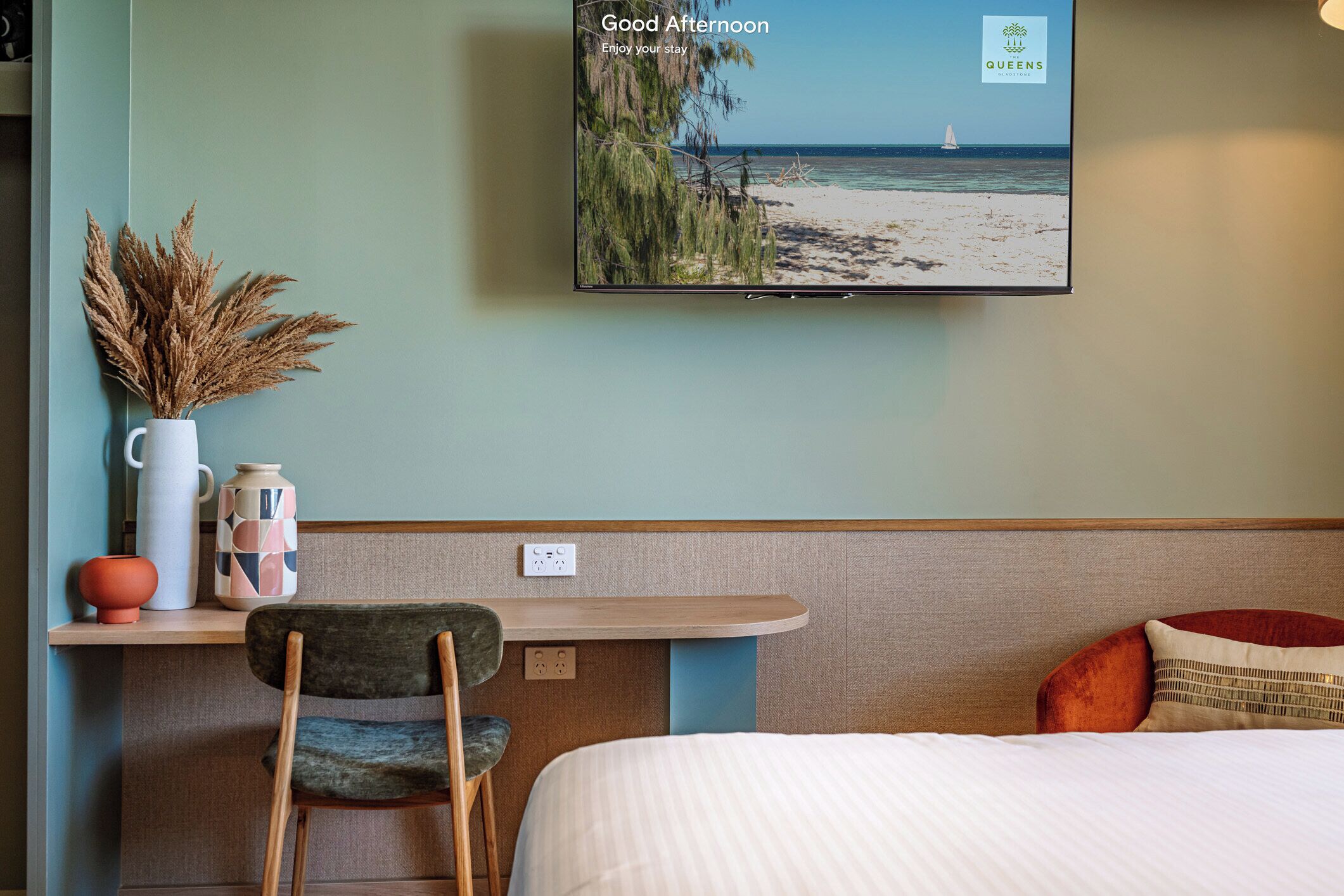 A hotel room with a bed, a desk with a chair, a TV displaying a beach scene, and a vase with dried plants.