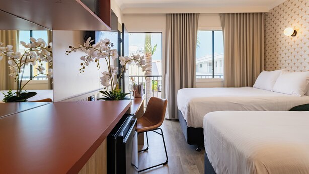 A hotel room with two beds, a desk with a chair, a large window with curtains, and a potted orchid plant. - The Queens Hotel Gladstone (Gladstone)