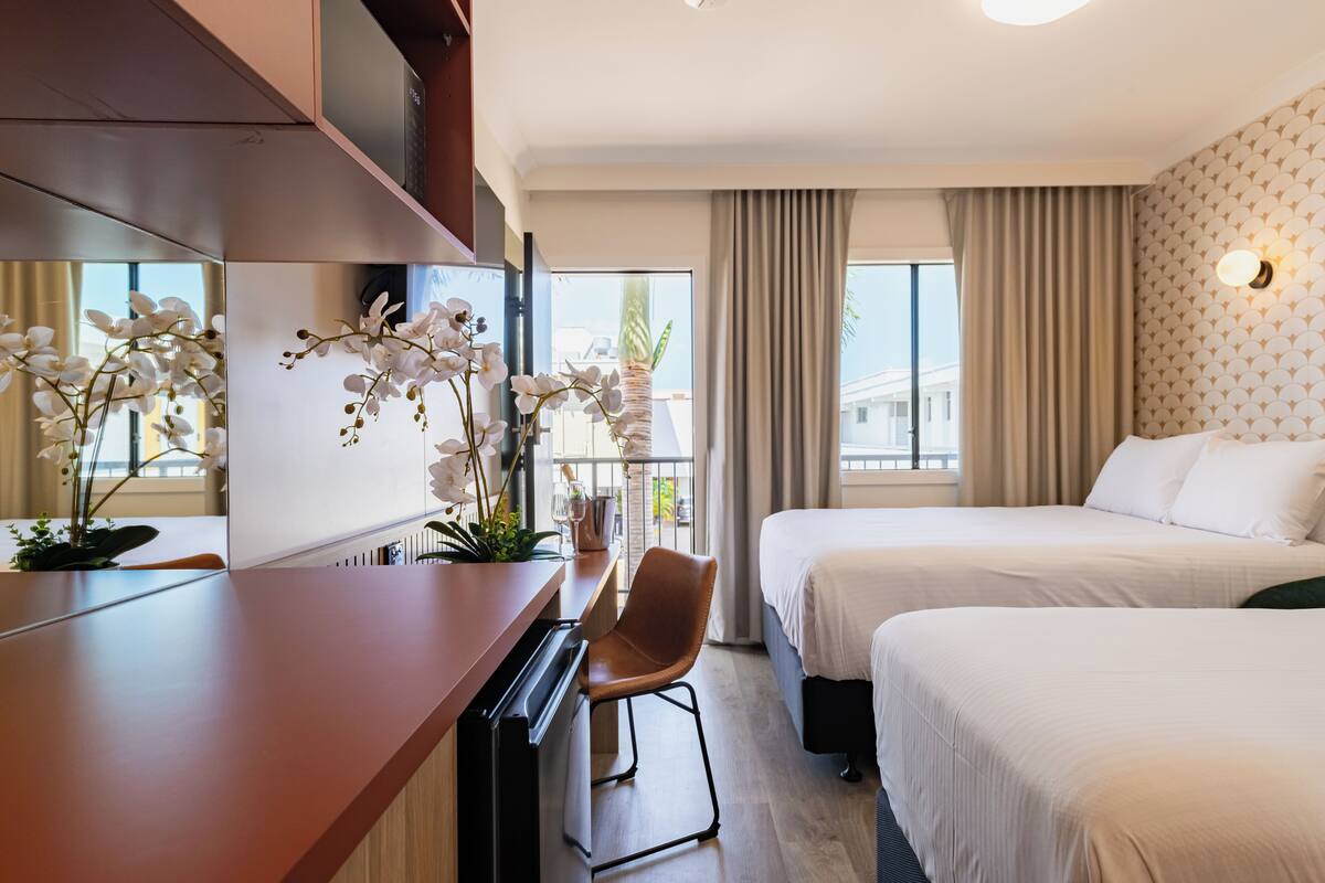 Ein Hotelzimmer mit zwei Betten, einem Schreibtisch mit Stuhl, einem großen Fenster mit Vorhängen und einer Orchidee im Topf.