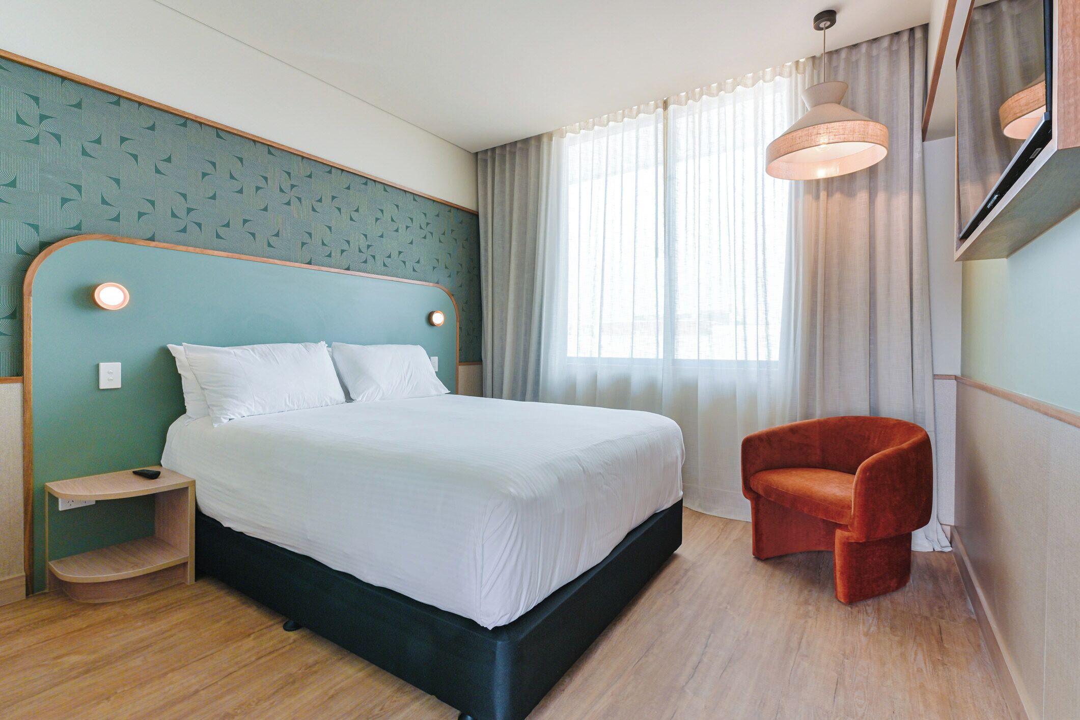 Ein modernes Hotelzimmer mit einem großen Bett, einem kleinen Nachttisch, einem roten Sessel und einem großen Fenster mit Vorhängen.