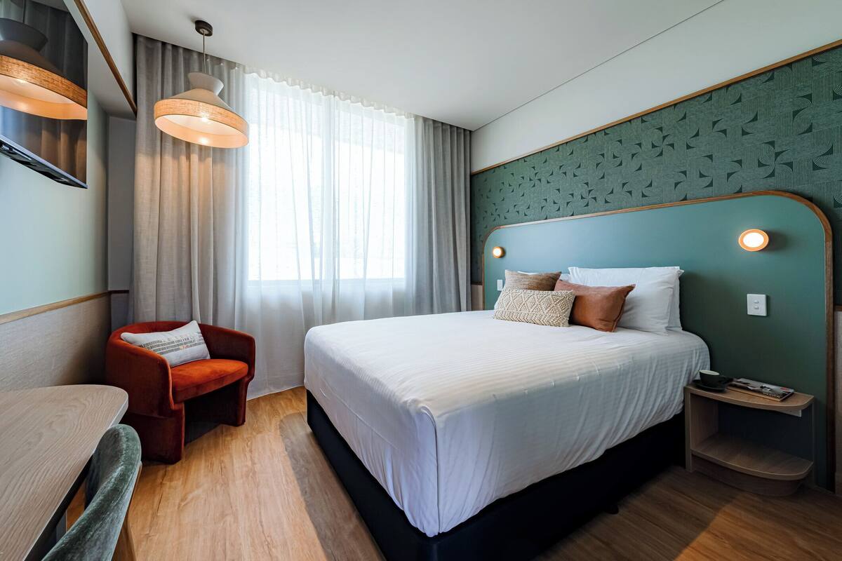 Ein modernes Hotelzimmer mit einem großen Bett, einem roten Sessel, einem Holztisch und Blick auf die Stadt durch große Fenster.