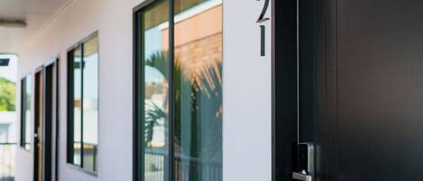 A modern building exterior with a black door, numbered 21, and a glass window reflecting a palm tree and building.
