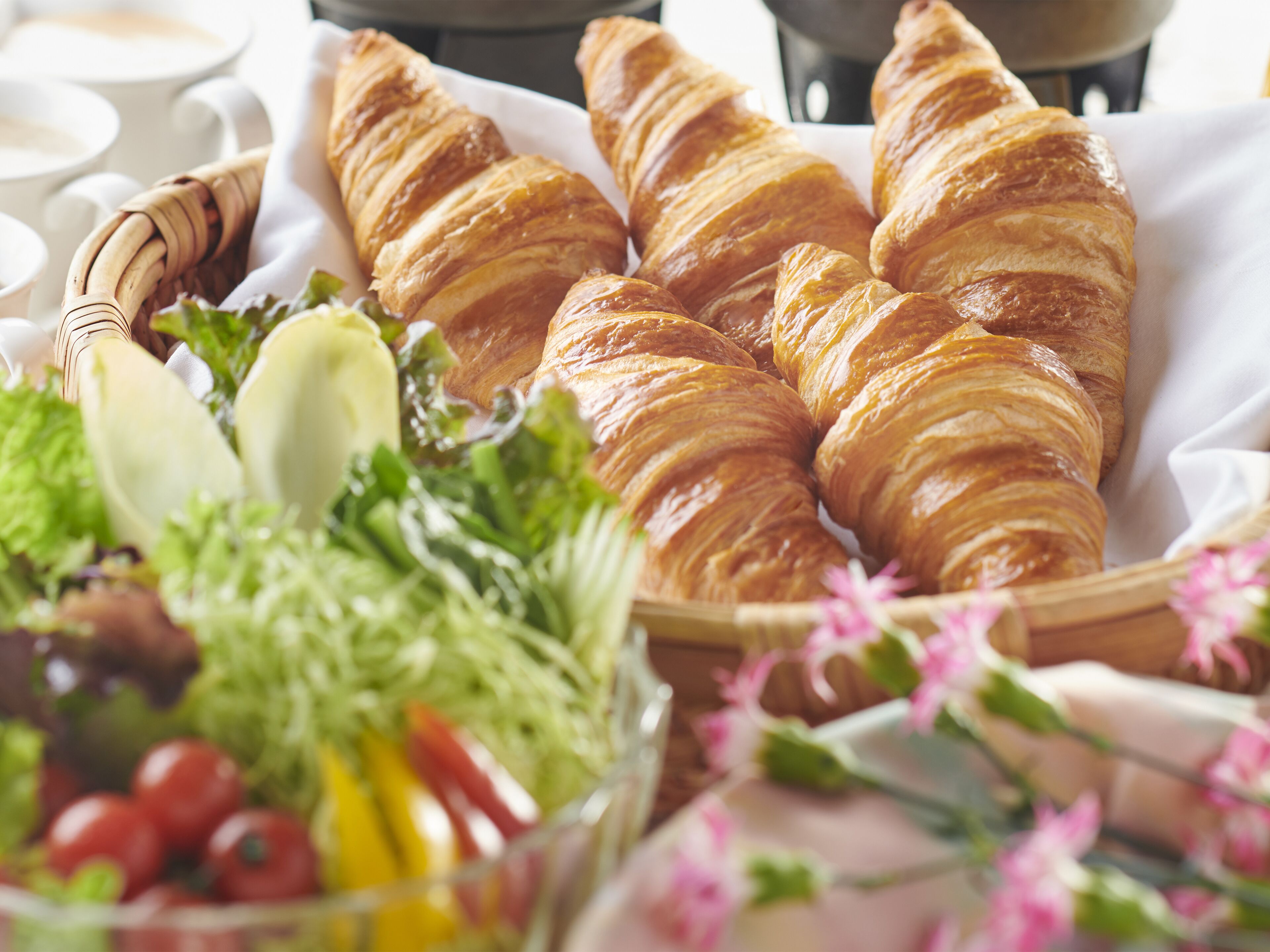 A basket of croissants on a table with a salad and flowers.