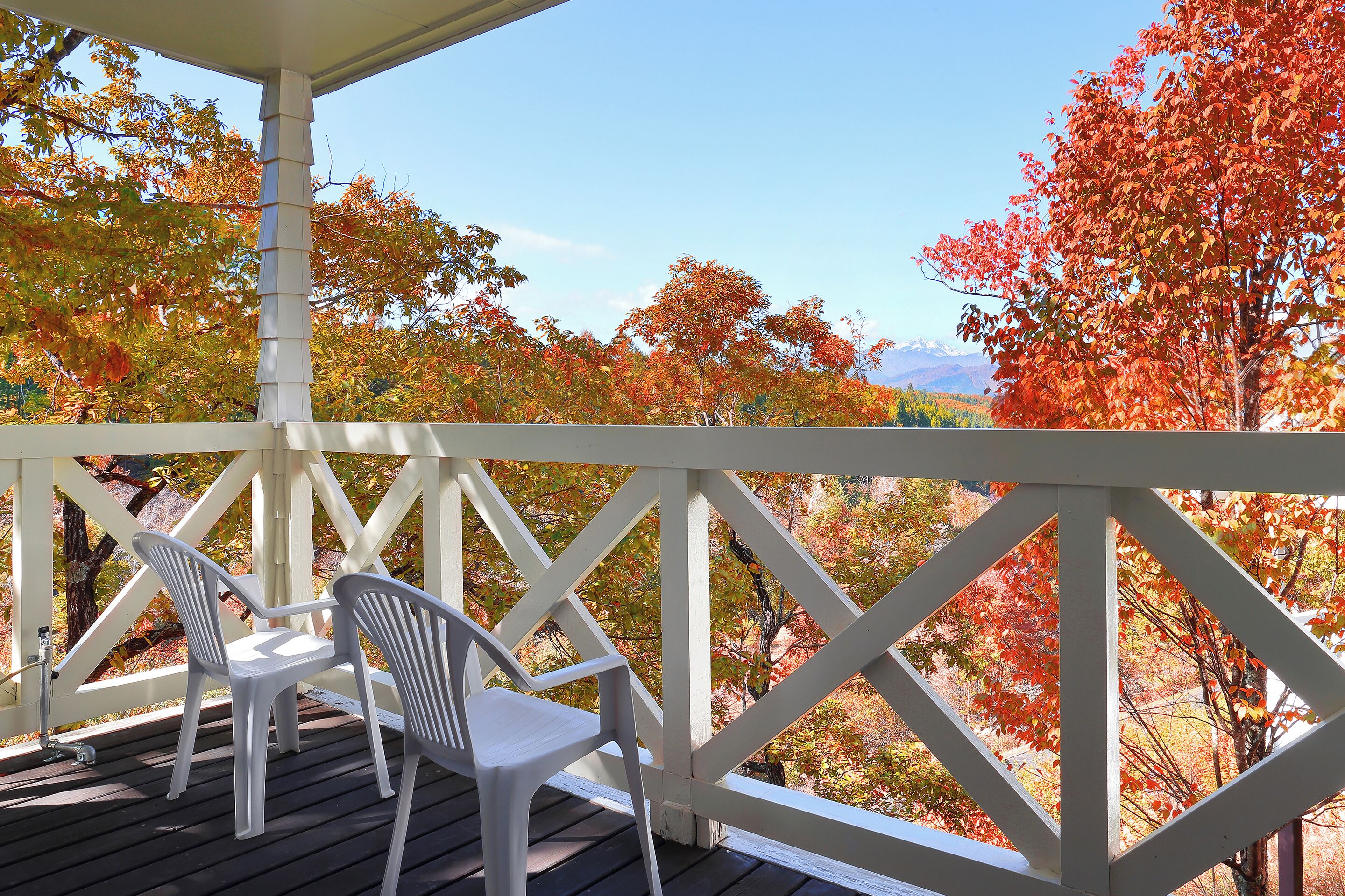 A covered outdoor area with a white railing, two white plastic chairs, and a view of autumn trees.