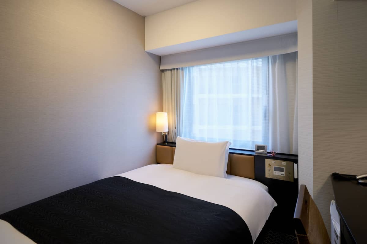 Une chambre d’hôtel équipée d’un lit, d’une table de chevet, d’une lampe, d’une fenêtre avec des rideaux et d’un thermostat.