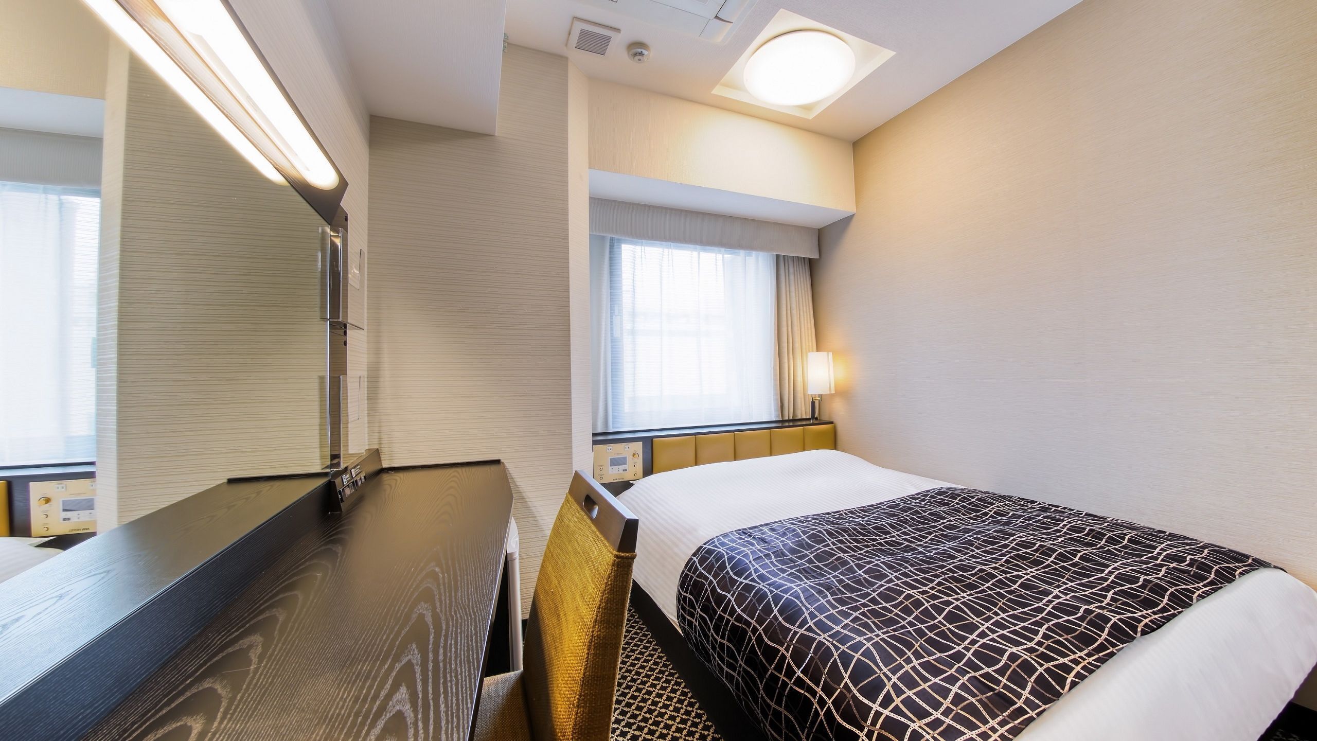 Une chambre d’hôtel comprenant un lit, une table de chevet, un miroir, une fenêtre avec des rideaux et un luminaire fixé au mur.