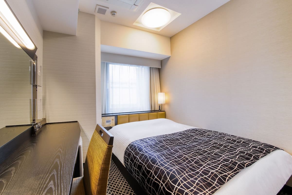 Une chambre d’hôtel comprenant un lit, une table de chevet, un miroir, une fenêtre avec des rideaux et un luminaire fixé au mur.