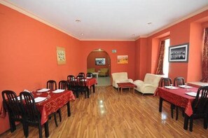 A dining area with wooden flooring and red walls, featuring tables set for dining and a sofa in the background.