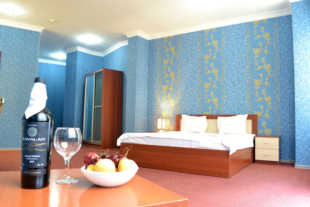 Une chambre d’hôtel avec un lit en bois, une table de chevet sur laquelle se trouvent une bouteille et un verre, et un bol de fruits.