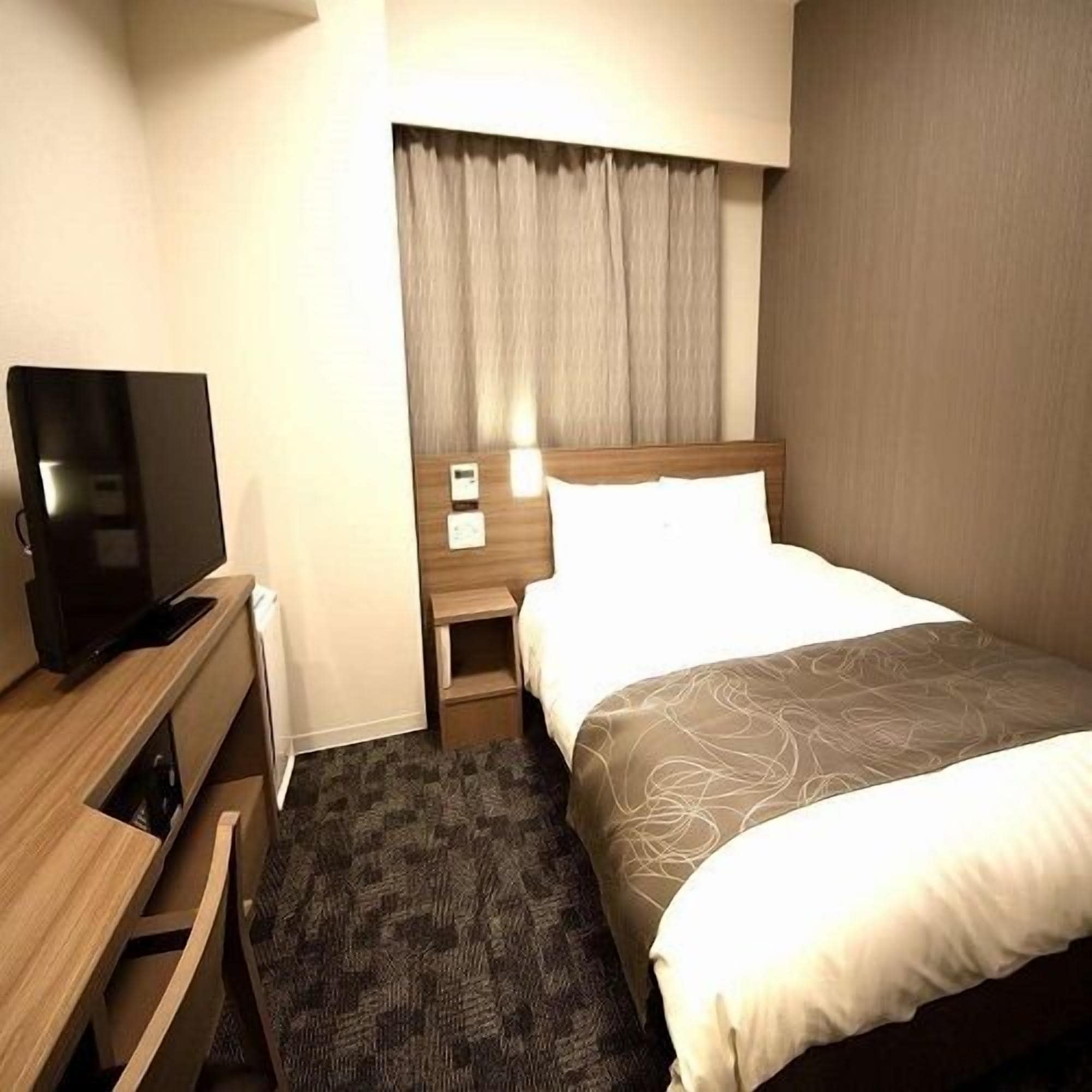 Quarto de hotel com uma cama, uma televisão sobre uma cômoda de madeira, uma mesa de cabeceira com abajur e uma janela com cortinas.