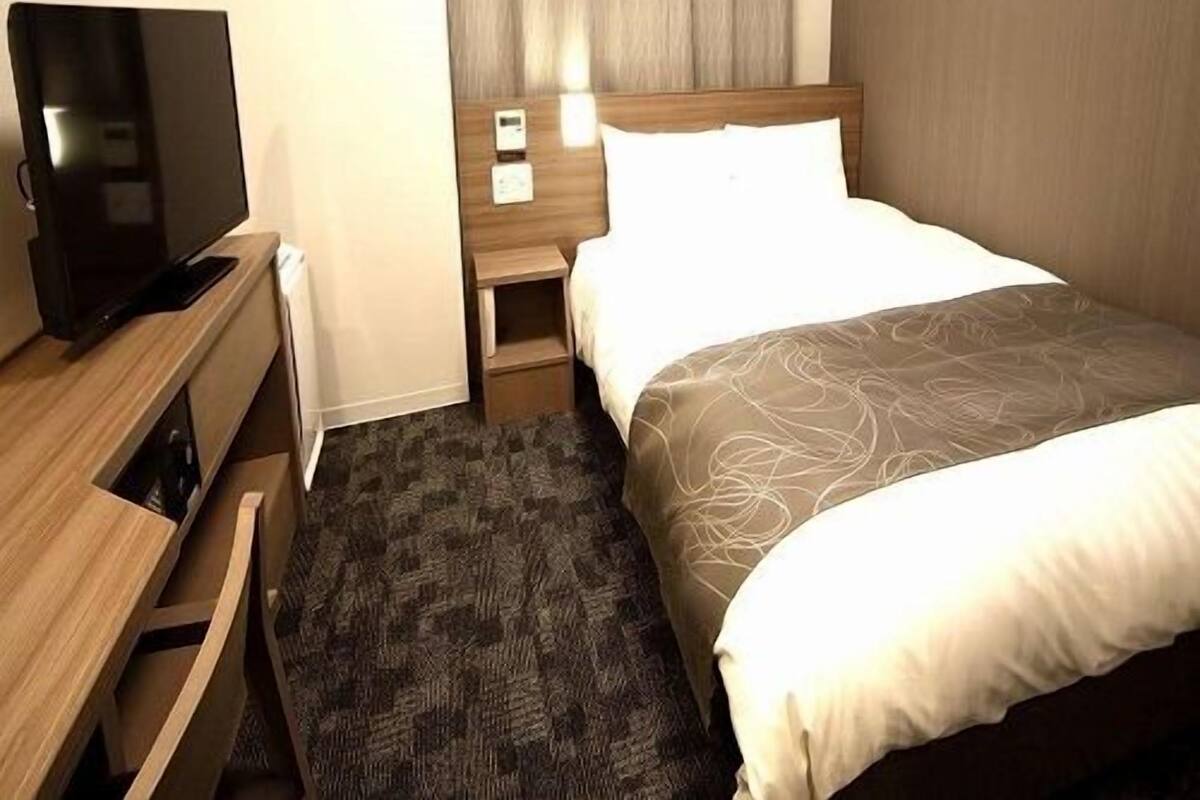 A hotel room with a bed, a TV on a wooden dresser, a nightstand with a lamp, and a window with curtains.