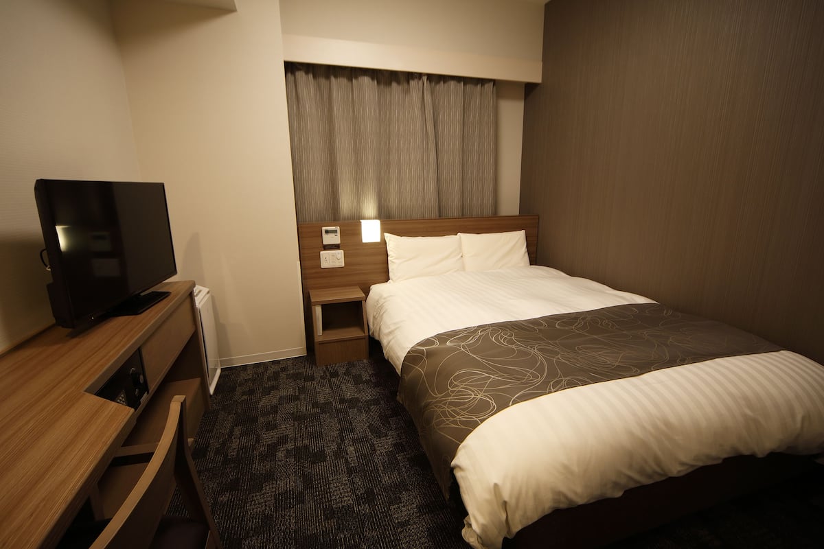 A hotel room with a bed, a desk with a television, a nightstand, and a window with curtains.