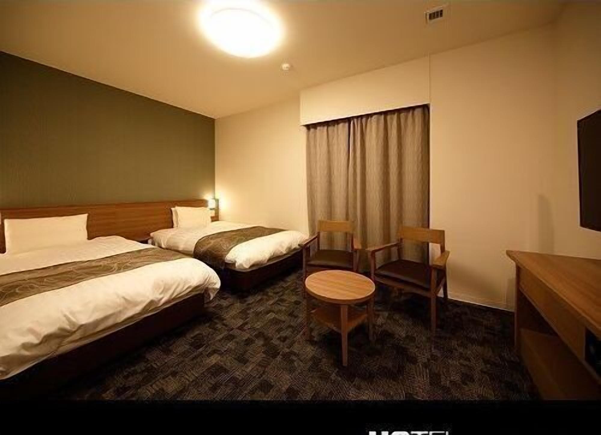 Quarto de hotel com duas camas, uma mesa redonda pequena, duas cadeiras de madeira, uma escrivaninha e uma janela grande com cortinas.